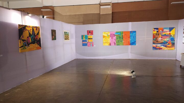 An art gallery room with various colorful paintings displayed on white partition walls. A spotlight is positioned on the floor in the center of the room, illuminating the artwork. Reminiscent of Shantiniketan's Kala Bhavana, the room boasts a wooden floor and high ceiling, exuding a Fine Arts College aura.