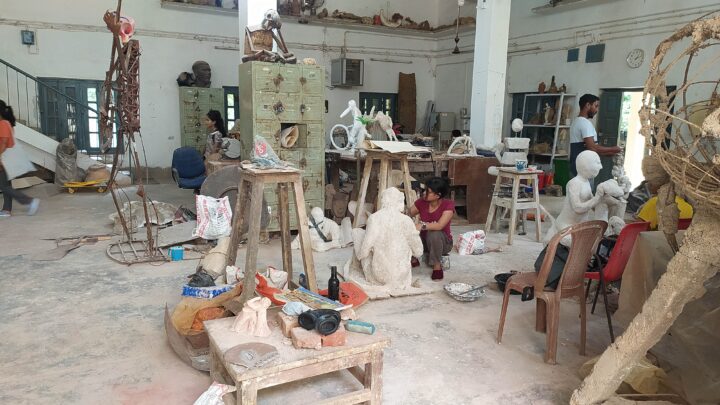 A spacious art studio reminiscent of the Kala Bhavana at Fine Arts College in Shantiniketan is depicted. Sacks, tools, and materials are scattered around the room. People are engaged in their work. Sculptures of human figures can be seen in different stages of completion.