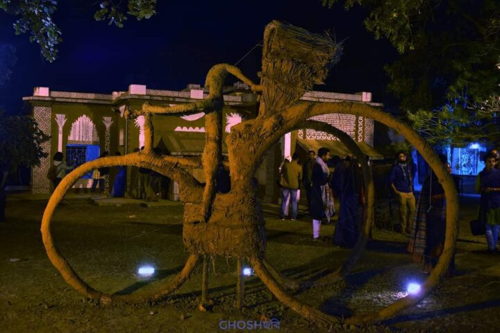 A large straw sculpture of a person riding a bicycle is displayed outdoors at night at Kala Bhavana, the Institute of Fine Arts in Shantiniketan. Illuminated with spotlights and ambient lighting, the scene features a building and people in the background, surrounded by trees and foliage.