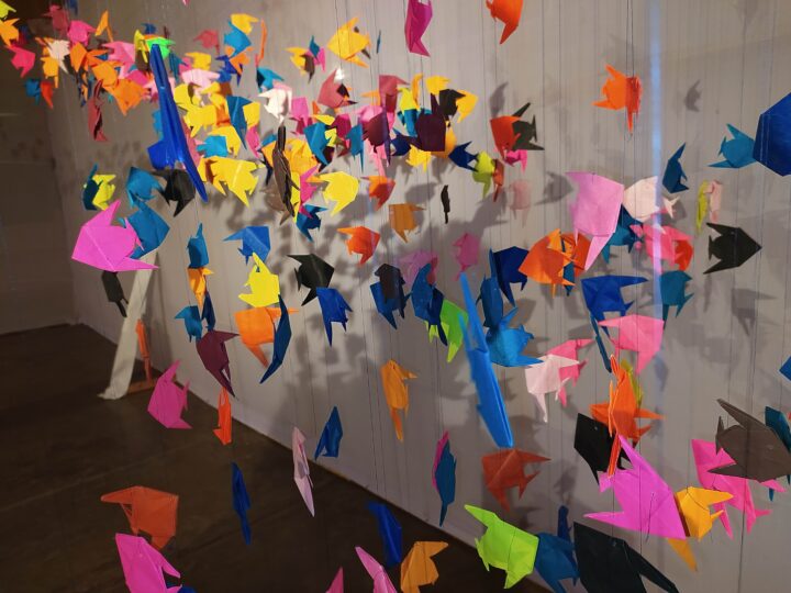 The image showcases an art installation from Kala Bhavana at Fine Arts College, Shantiniketan, featuring numerous colorful origami birds hanging from strings. The birds are arranged in a suspended manner, creating a visually dynamic and vibrant display against a white background.