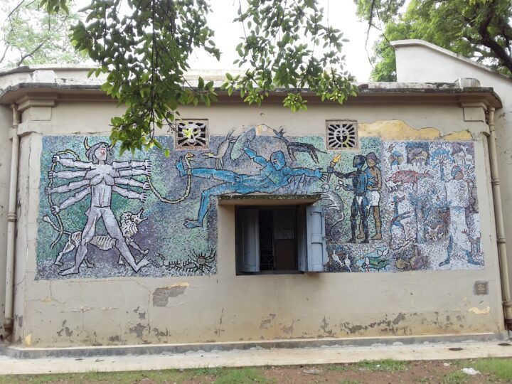 A mural covers the exterior wall of a building in Shantiniketan. The artwork, reminiscent of the Fine Arts taught at Kala Bhavana, features various human figures, including one with multiple arms and another in blue, amidst abstract designs. The building has light peeling paint, two small ornamental windows, and a partially open window at the center.