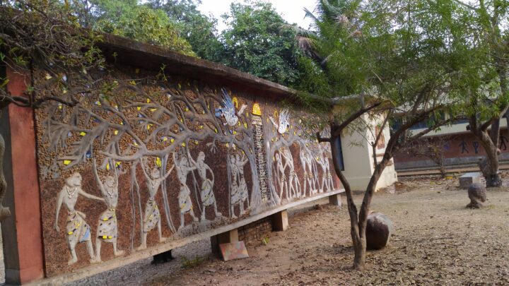 A large outdoor mural, reminiscent of the artistic style seen at Shantiniketan's Kala Bhavana, depicts human figures and trees created with mosaic tiles and various materials. Mounted on a long wall with nearby vegetation, a smaller stone object sits in front, adding to the evocative scene.