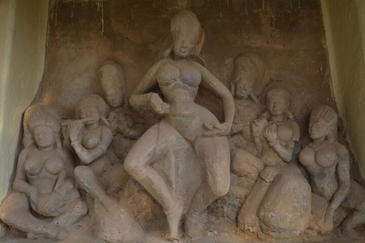 A stone carving at Kala Bhavana in Shantiniketan depicts a central female figure in a dynamic pose, surrounded by five other figures, all with detailed and expressive features. The scene is carved into a recessed area in a wall, with weathered and aged texture evident on the stone surface.
