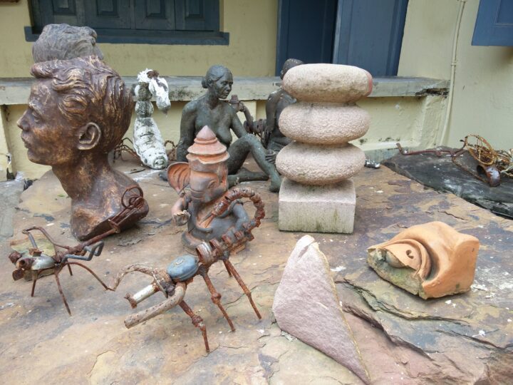 An assortment of fine arts sculptures and artistic pieces is displayed on a stone surface. The collection includes human busts, abstract forms, animal figures, and other intricate models made from various materials. The background includes a beige wall with blue window frames reminiscent of Shantiniketan's Kala Bhavana.