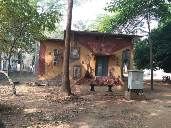 A small building with an artistic mural on its exterior wall. The mural, reminiscent of Shantiniketan's famed Kala Bhavana, depicts various objects including a bicycle, a window with bars, and a clock. Tall trees surround the building, and a white electrical box stands nearby. The ground is covered with dry leaves.