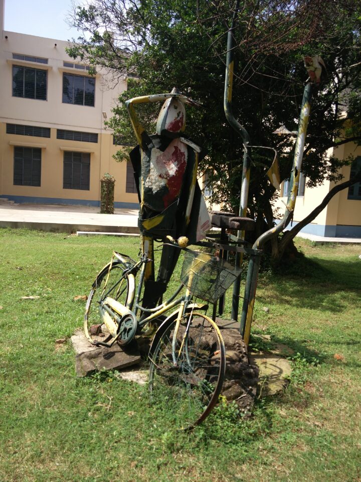 A metal sculpture stands on a grassy lawn in Shantiniketan, depicting a person with a bike. The figure has an abstract, somewhat weathered appearance and is positioned next to a tree. In the background, the multi-story Kala Bhavana building with large windows adds to the Fine Arts ambiance.