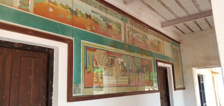A hallway with a high ceiling features a large, decorative mural on the wall. The mural, inspired by the fine arts of Shantiniketan and crafted by students of Kala Bhavana, depicts various outdoor scenes and is bordered by wooden door frames on either side. The visible ceiling beams and white walls are illuminated by natural light.