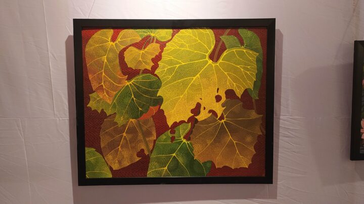 A framed artwork from the Fine Arts College at Kala Bhavana, Shantiniketan, features a mosaic of large, overlapping leaves in shades of green, yellow, and brown displayed on a white wall. The leaves have intricate vein patterns, creating a textured and vibrant composition.