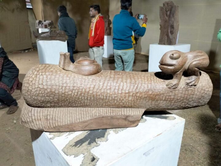 A wooden sculpture featuring a frog and snail on a textured log is displayed on a white pedestal. In the background, several people, reminiscent of visitors at Kala Bhavana in Shantiniketan's Fine Arts College, admire other sculptures in the gallery-like setting.