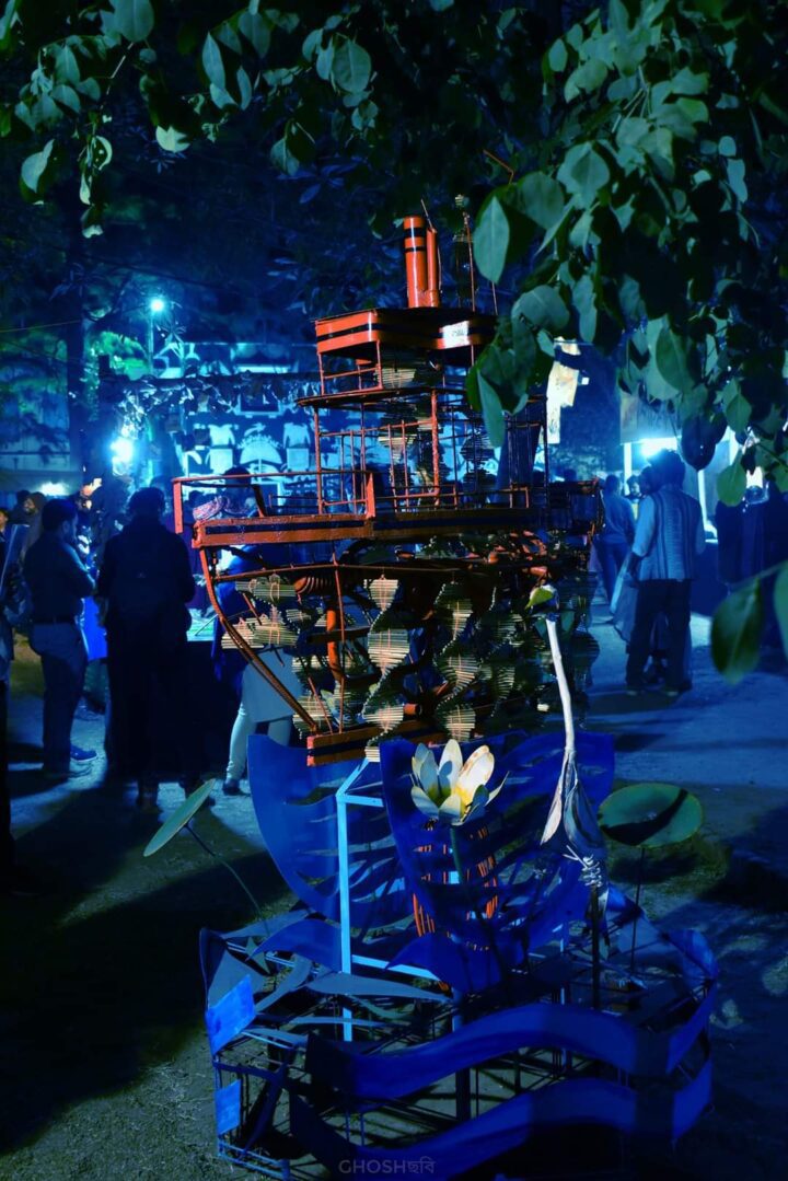An intricate red and blue model of a ship is displayed outdoors at night, surrounded by trees and various people in the background. The scene is illuminated by blue lighting, creating a vibrant and surreal atmosphere reminiscent of the artistic ambiance found at Kala Bhavana, Shantiniketan’s Institute of Fine Arts.