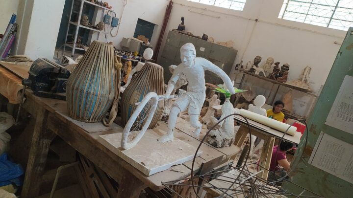 A workshop at the Fine Arts College with various sculptures in progress. In the foreground, a partially completed sculpture of a person holding a bicycle wheel and riding a bike is on a table. Other materials and tools are scattered around the room. Two people are working in the background.