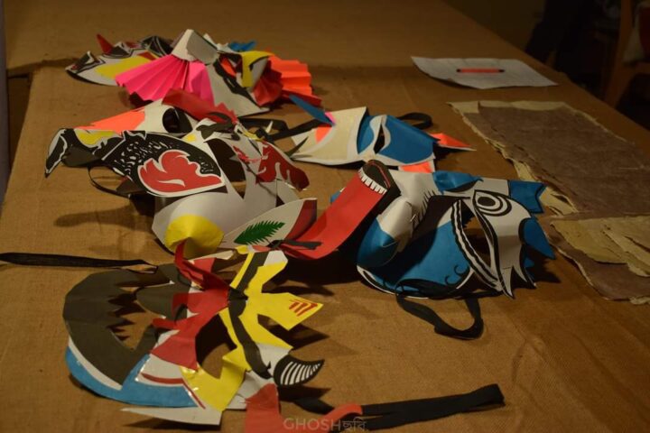 A table displays colorful, handmade masks crafted from paper. The creations, reminiscent of the vibrant artistry seen at Kala Bhavana in Shantiniketan, feature various designs including animal motifs and abstract shapes in red, yellow, blue, and black. Paper scraps and a sheet of paper are scattered around the table.