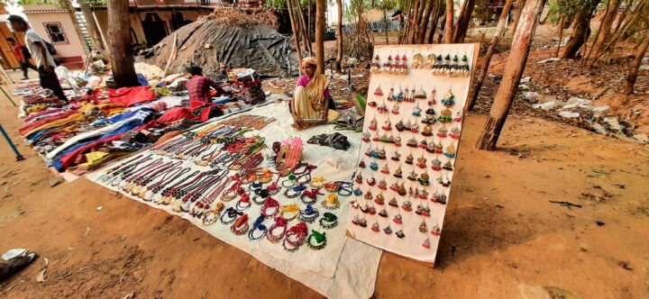 A woman sits behind a display of assorted jewelry and accessories laid out on a cloth, including necklaces and hairbands. Next to her is a board featuring rows of earrings. The background shows tall trees and people browsing.