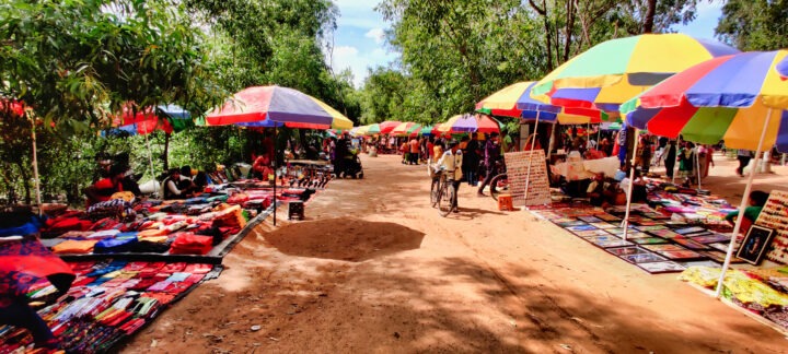 A vibrant outdoor market scene with colorful umbrellas shading various stalls. Vendors display textiles and framed artworks along a dirt pathway lined with trees. Shoppers browse the items, and a person on a bicycle rides through the market.