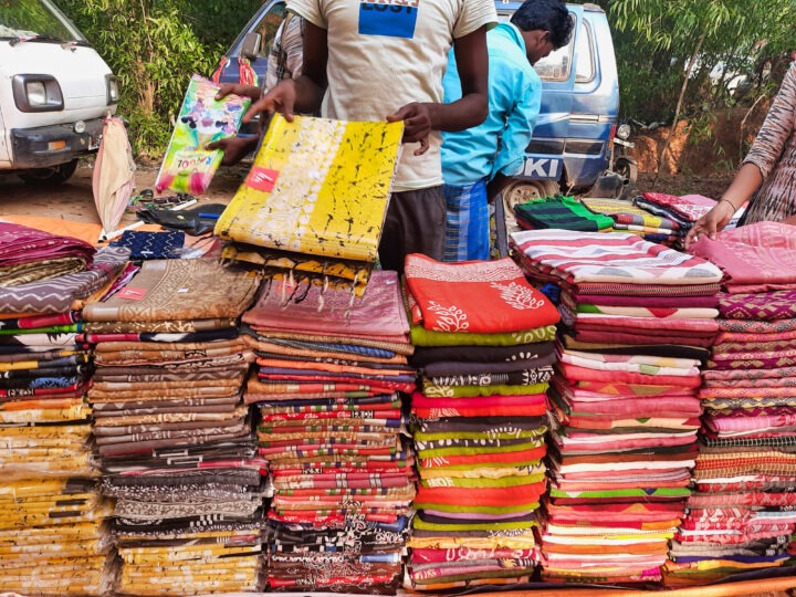 A street vendor is holding a colorful piece of fabric over a table stacked with various printed and patterned textiles. Two vehicles are in the background, and another person is partially visible standing nearby.