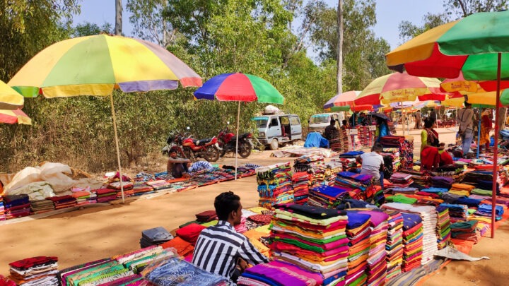 Outdoor market scene with vibrant umbrellas providing shade. Colorful stacks of fabric and clothing are displayed on ground stalls. Vendors and customers are visible among the items. Trees and parked vehicles can be seen in the background.