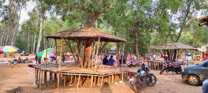 A market scene in a wooded area with wooden platforms and structures. People are gathered, and colorful umbrellas are scattered around. Motorbikes and a parked car are visible, with trees surrounding the area.