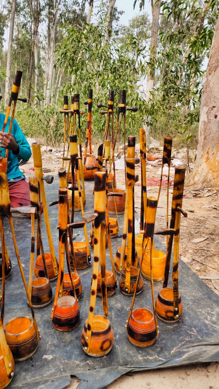 A collection of traditional stringed instruments displayed on a tarp outdoors. The instruments feature long necks and round bodies in shades of brown and orange. Trees and plants are visible in the background, with a person partially seen to the left.