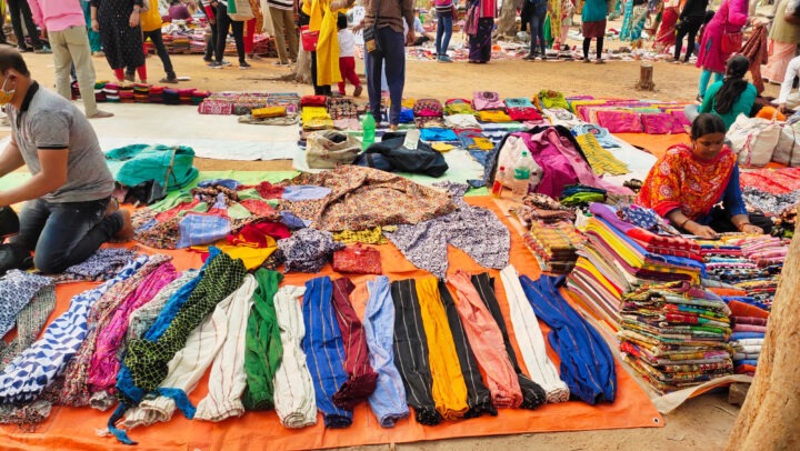 A busy outdoor market scene with vendors displaying colorful fabric and clothing items on the ground. Shoppers are walking around, examining the goods. Brightly colored textiles, bags, and scarves are spread out on orange and white sheets.