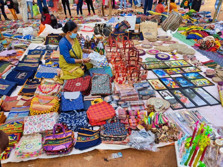 A vendor in a yellow saree arranges handmade bags, pouches, and woven crafts displayed on a sheet at an outdoor market. People are browsing nearby. The scene is vibrant with colorful textiles and artisanal items.