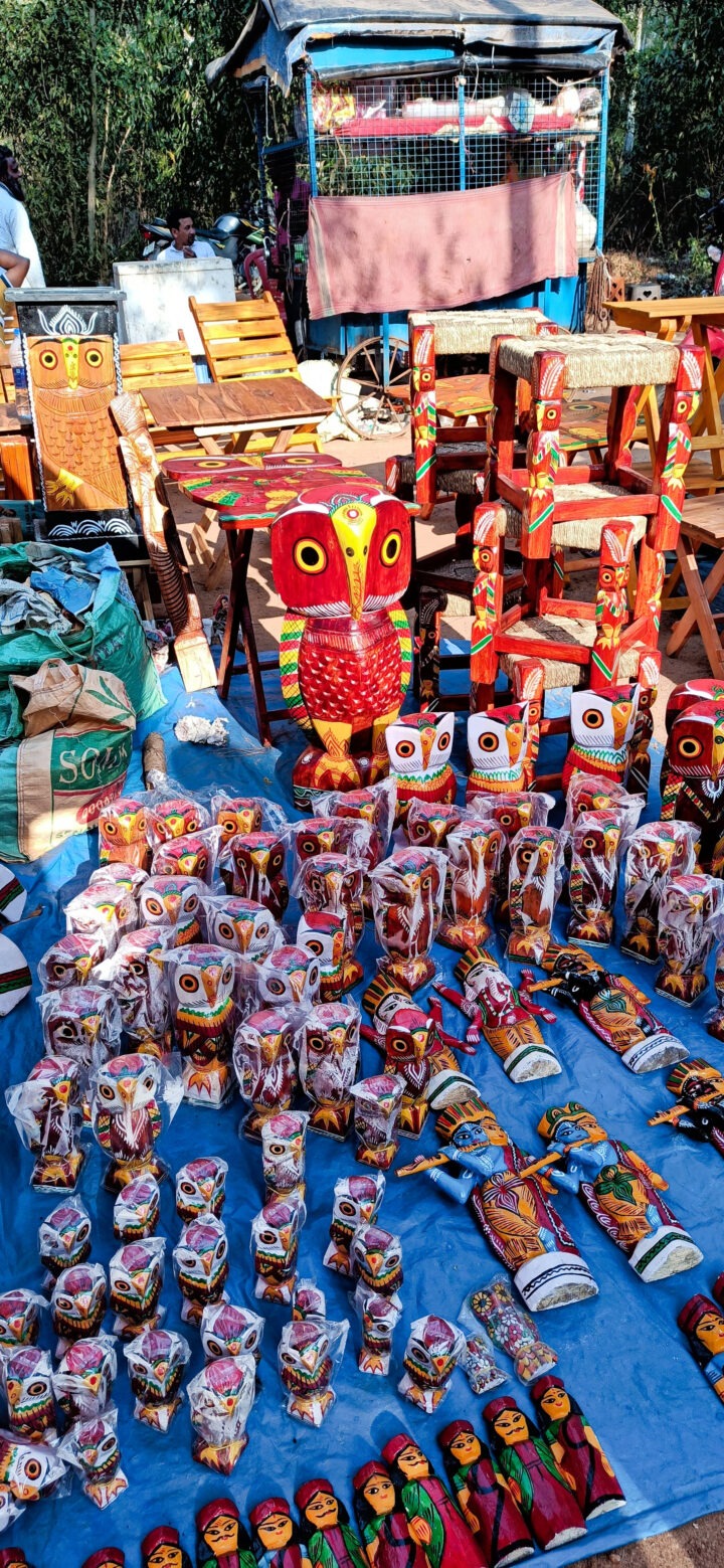 A marketplace stall displays colorful owl-themed handcrafted items, including wooden sculptures and dolls, arranged on a blue cloth. In the background are wooden furniture pieces. The setup is outdoors on a sunny day.