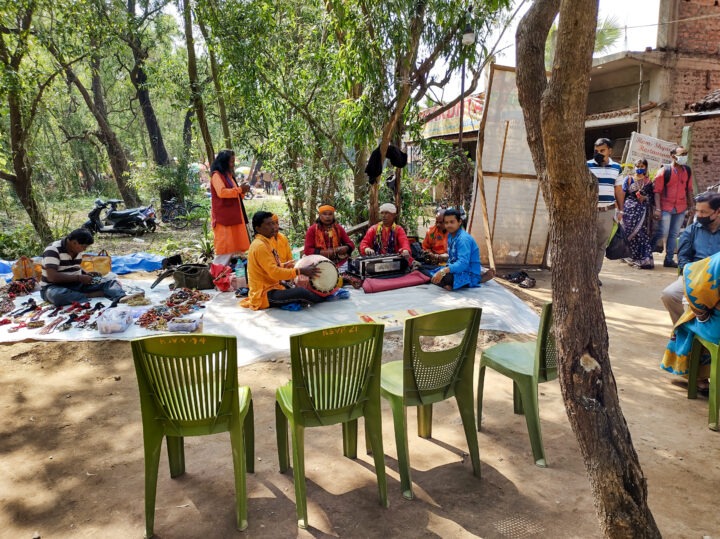 A group of musicians sits on a mat playing traditional instruments outdoors, surrounded by trees. Chairs are set up nearby, and various crafts are displayed on the ground. People stand in the background, some wearing masks.