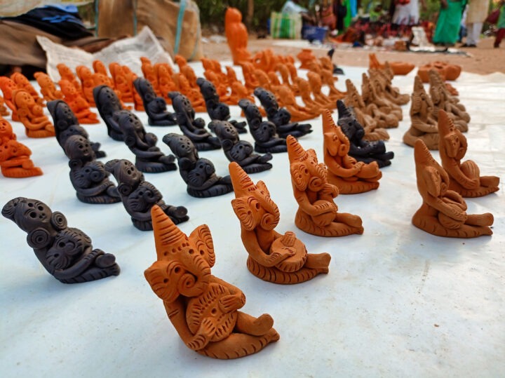 Rows of small clay figurines are displayed on a white cloth. The figures, in shades of brown and black, have intricate designs and are placed in neat lines. People and greenery are visible in the background.