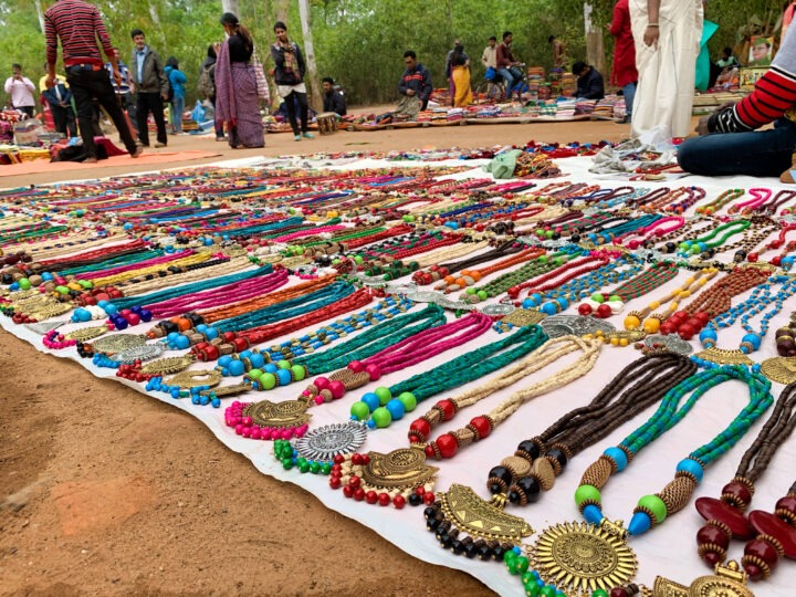 Rows of colorful, assorted beaded necklaces and jewelry displayed on white cloth at an outdoor market. People browse in the background, set against a backdrop of trees.