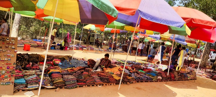 A market stall with colorful umbrellas offers various fabrics and textiles displayed on the ground. In the background, people browse and shop in a shaded outdoor area. Accessories like earrings hang on a display board.