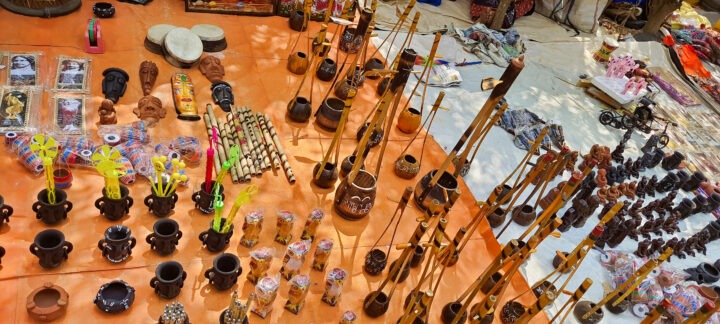 A display of various handcrafted items on an orange cloth. The items include wooden flutes, decorated cups, small clay figures, and assorted trinkets. The setup is part of an outdoor market or craft fair.