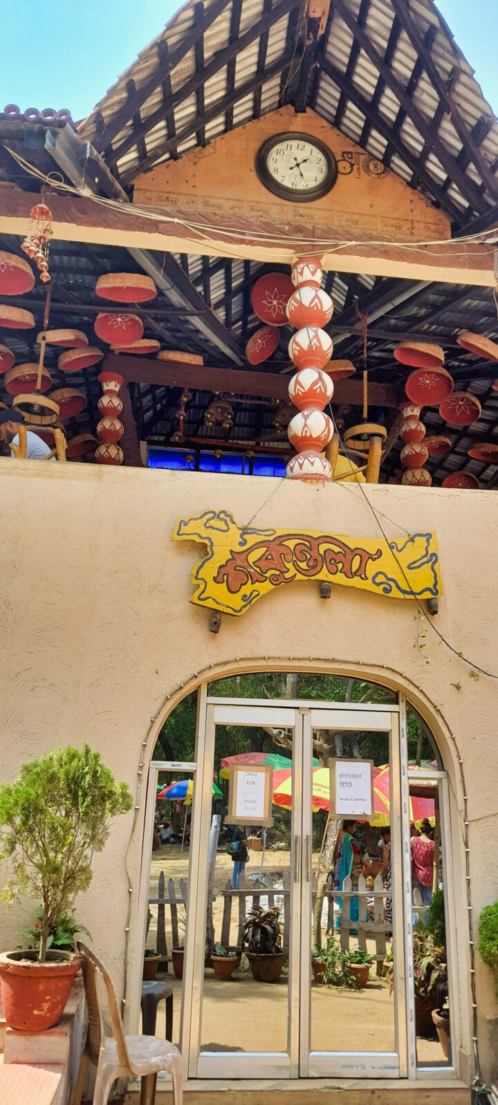 The image shows the entrance of a building with an arched doorway and a yellow sign above it. The upper floor has a wooden roof with hanging pots and a clock. People are visible inside through the glass doors. A potted plant is in front.
