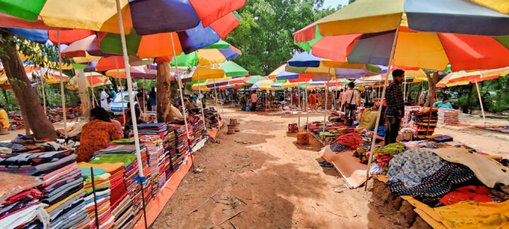 A vibrant outdoor market scene with colorful umbrellas providing shade. Various stalls display stacks of fabrics and clothes. People are browsing the merchandise under the lush green trees on a sunny day.