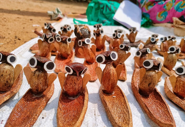 Handcrafted owl figurines made from natural materials are arranged on a white textured surface. Each figurine features large, round eyes and is set on a wooden base. Bright sunlight casts shadows, and colorful fabric is visible in the background.