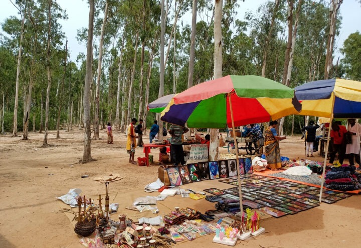 An outdoor market in a forested area features colorful umbrellas shading various stalls. Displayed items include artwork, crafts, and souvenirs. People are browsing and engaging with vendors amidst tall trees in the background.