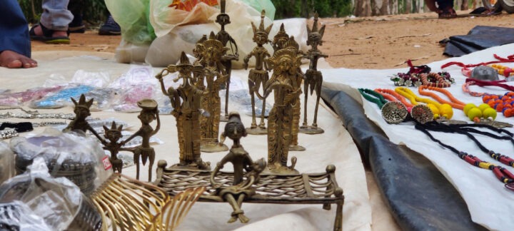 A variety of handcrafted metal figurines are displayed on a white cloth at an outdoor market. The figures depict musicians and seated individuals. Decorative jewelry and assorted items are placed nearby on the ground.