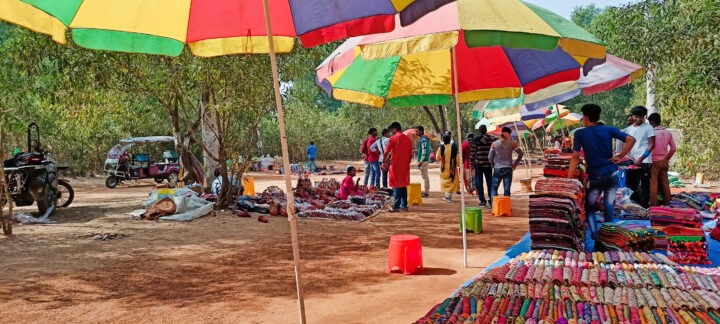 Outdoor market scene with colorful umbrellas providing shade. Stalls display various textiles and crafts on tables. People walk around, browsing and shopping. Trees are visible in the background, and the ground is sandy.