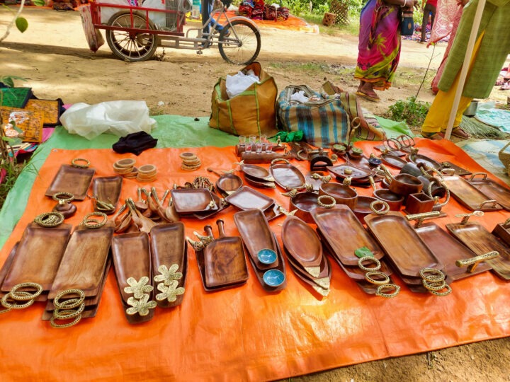 A variety of wooden trays, dishes, and decorative items are displayed on an orange sheet at an outdoor market. A bicycle and people are in the background, with colorful fabrics and bags visible nearby.