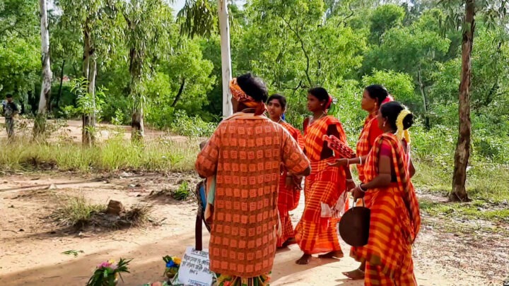 A group of people dressed in traditional attire gather outdoors, surrounded by lush green trees. Some are holding musical instruments. The setting appears to be a rural area with a dirt pathway and scattered plants.