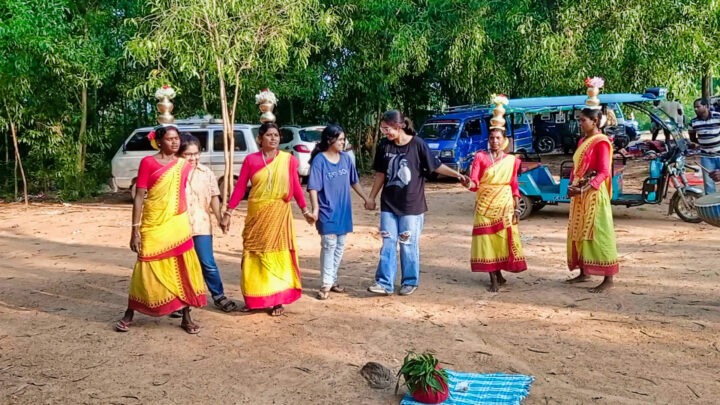 A group of women in yellow and red traditional attire perform a dance outdoors, with pots on their heads. Two people in casual clothes stand among them. Blue vehicles and green trees are in the background. A mat and some items are on the ground.