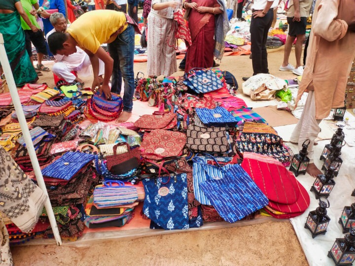A busy outdoor market stall displays a variety of colorful fabric bags with different patterns. Several people are browsing and examining the items. In the background, more shoppers and stalls are visible.