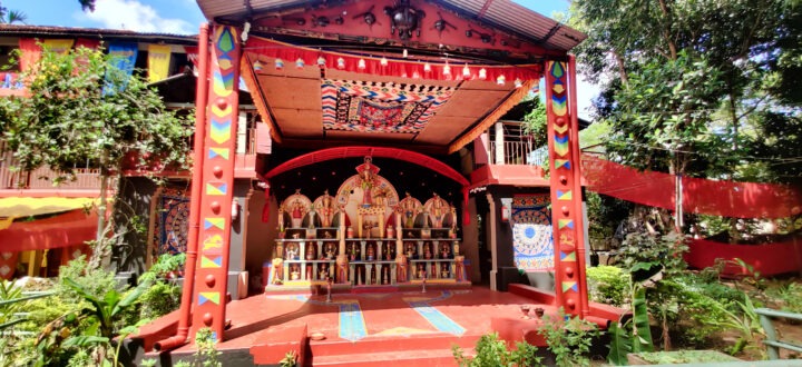 A colorful outdoor stage with vibrant decorations featuring intricate patterns and motifs. The backdrop displays a row of ornate structures, surrounded by lush greenery and set against a clear blue sky.