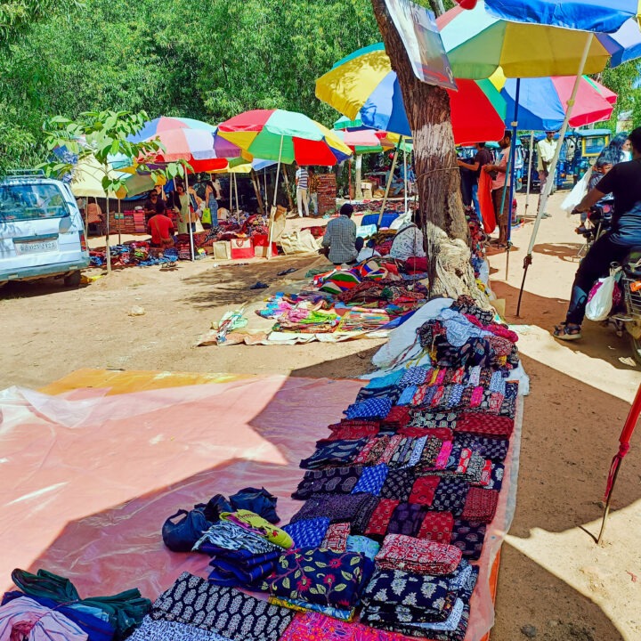 A vibrant outdoor market scene with colorful fabrics displayed on tables. Multiple umbrellas provide shade, and vendors and shoppers are scattered around. Trees and parked cars are visible in the background.