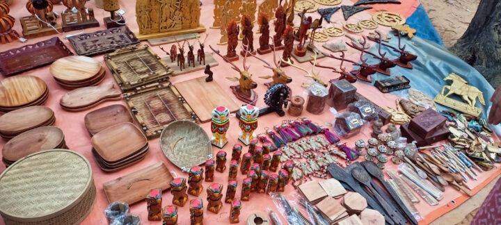 A table is covered with various handcrafted wooden items, including dishes, figurines, small statues, and decorative pieces. There are also woven baskets, colorful masks, and carved animals, all displayed on a cloth-covered surface.