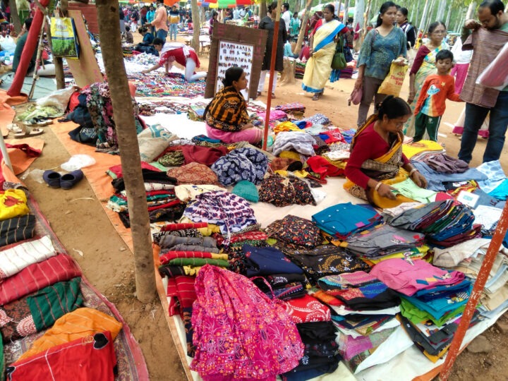 A bustling outdoor market featuring vendors displaying colorful fabrics, clothes, and textiles on the ground. Shoppers walk and browse the selections. Trees are scattered throughout the area, and a few signs are visible.