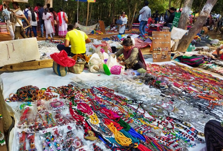 Outdoor market scene with vendors displaying colorful jewelry and crafts on white sheets. People browse items like necklaces, bracelets, and earrings. Trees and more shoppers are visible in the background.