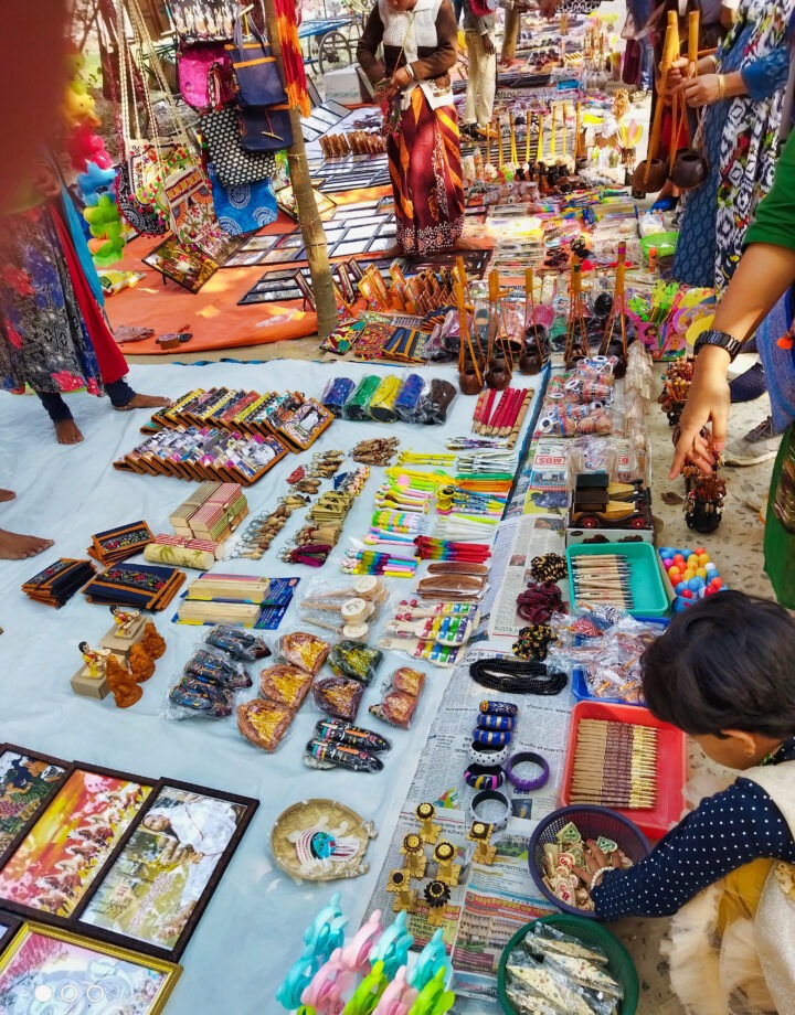 A vibrant market stall displays an array of colorful items, including jewelry, pens, wooden crafts, and decorative goods, spread out on a white sheet. Shoppers are browsing and selecting items. Sunlight casts shadows across the scene.