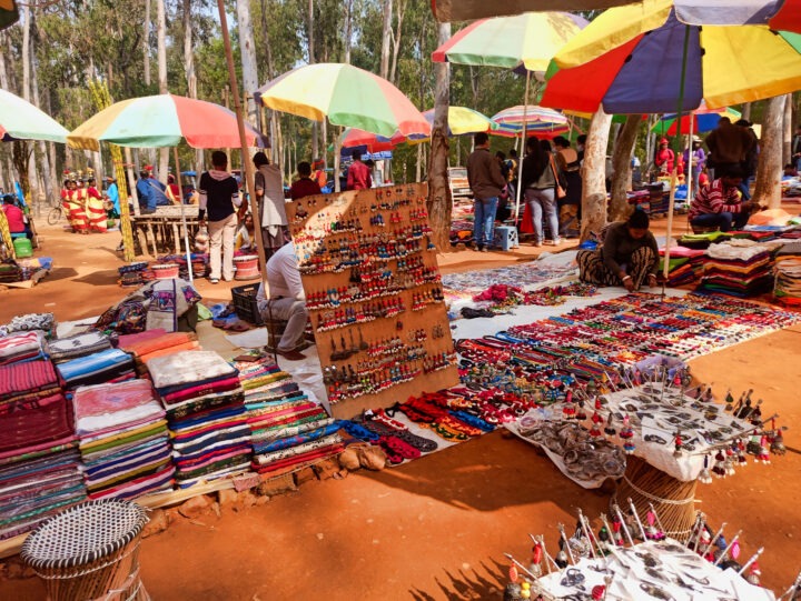 A vibrant outdoor market on a sunny day features colorful umbrellas, stacks of fabric, and displays of hand-crafted jewelry. People browse and shop among the stalls, with trees in the background providing natural shade.