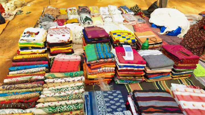 A display of colorful fabrics and garments arranged on the ground, featuring various patterns and textures. The fabrics are stacked in neat piles, creating a vibrant and organized market stall setting.