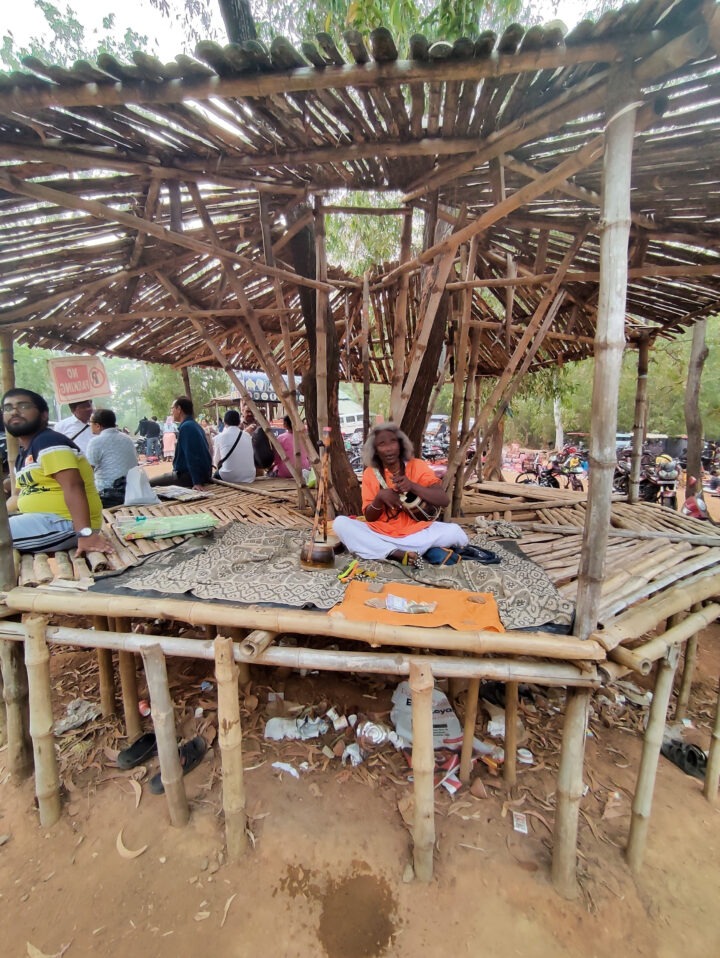 A person wearing an orange top and white pants is seated on a bamboo platform, playing a flute. People are seated around the structure, and trees are visible in the background. The ground is covered with dirt and litter.