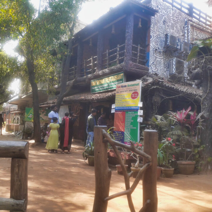 A rustic restaurant with a wooden facade and a sign reading "Dhaba" is surrounded by trees. Several people stand outside, along with a menu board featuring colorful options. The ground is covered with reddish dirt.